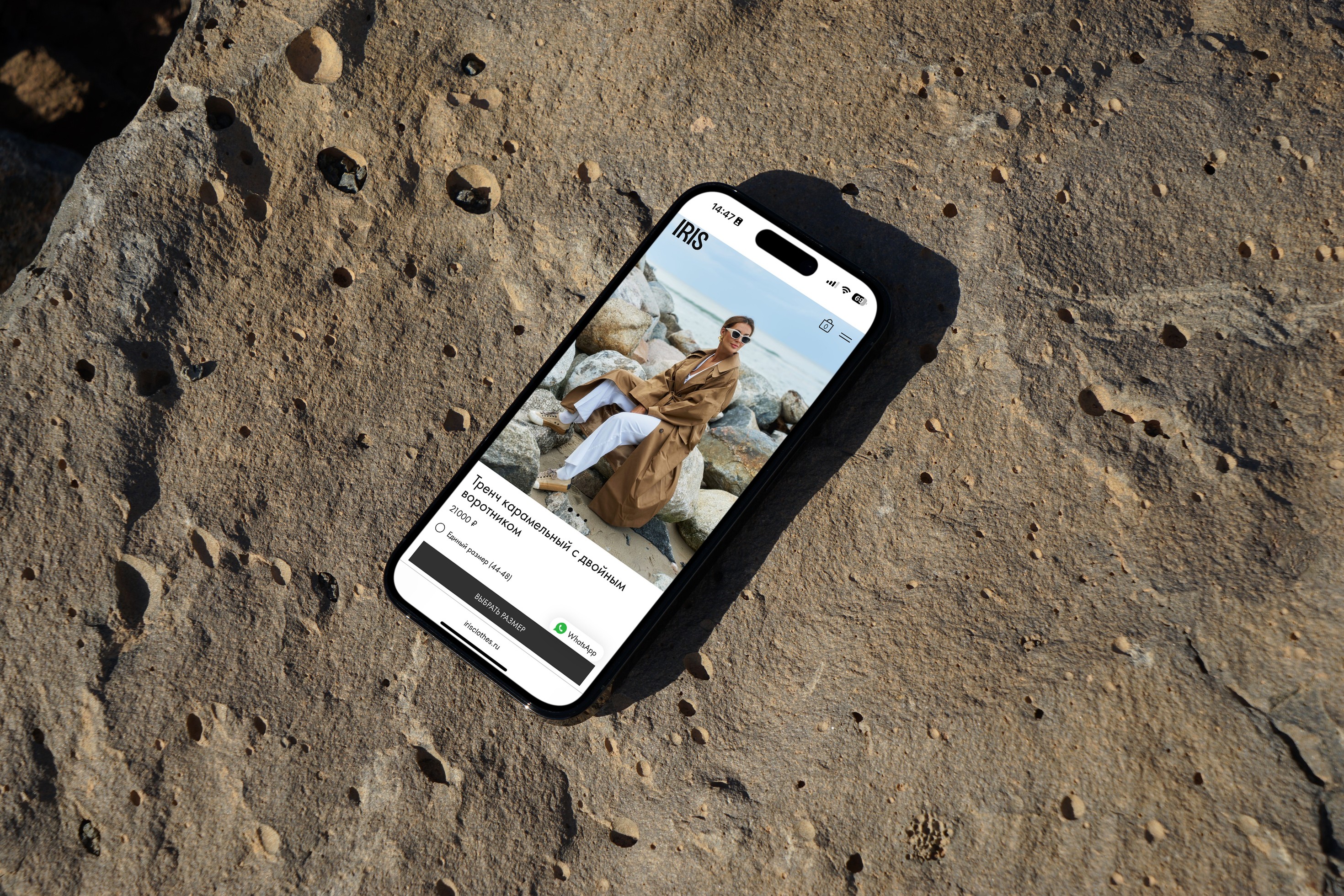 Мобильный телефон лежащей га камне с открытой в браузере страницей карточки товара пальто с указанием цены, описания и кнопкой «Добавить в корзину»